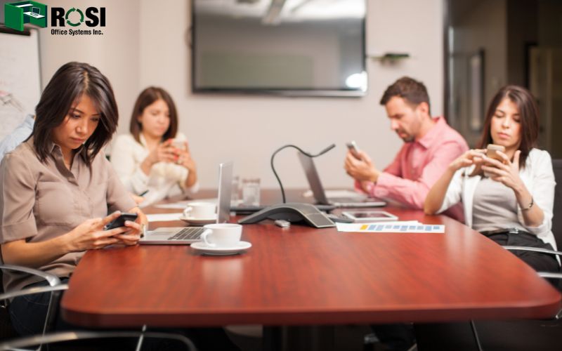 A group of people sitting in a meeting room but being distracted by their phones