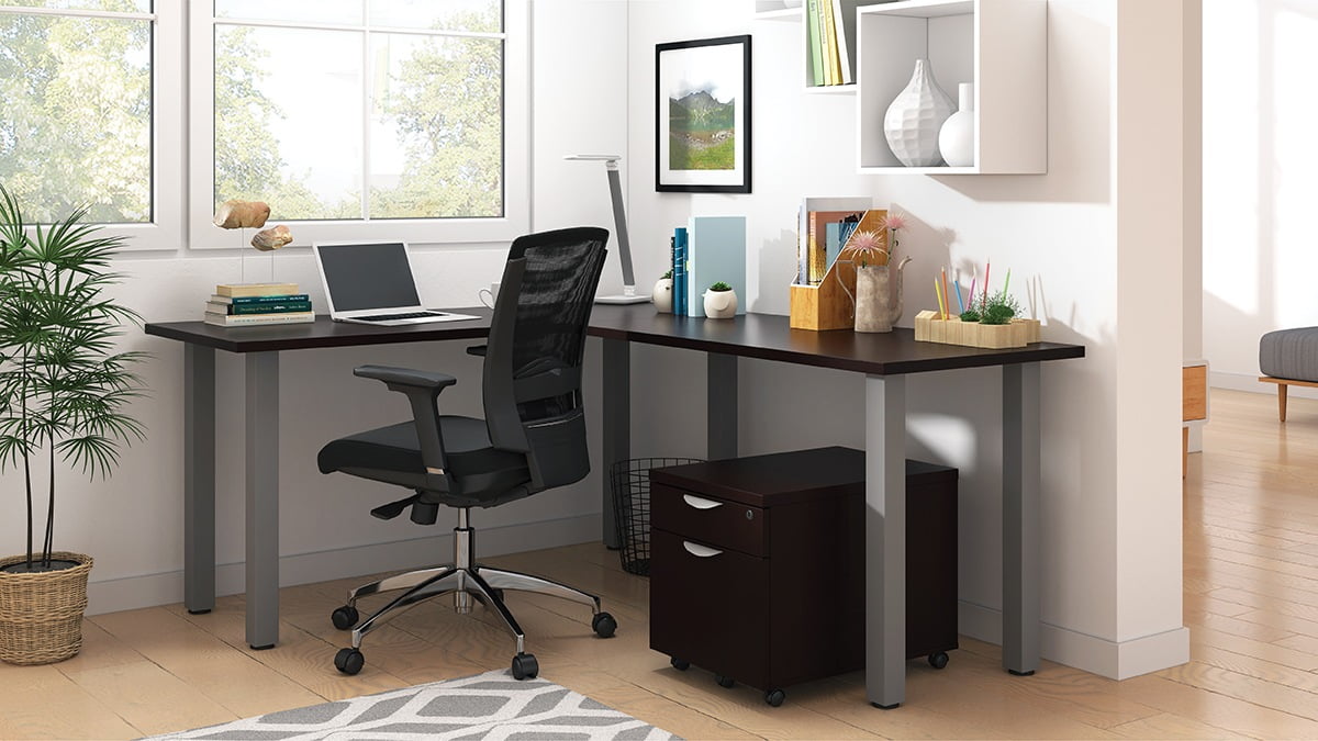 Studio shot of OTG's Home Office, with an L-shaped desk with a tungsten metal frame. The surface is finished in American Mahogany. A matching set of drawers has been rolled under the desk, at the left of the mesh back chair. To the left of the laptop is a set of books and an OTG LED Desk lamp in the corner.