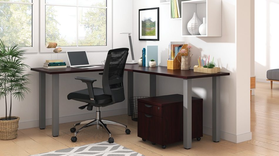 Studio shot of OTG's Home Office, with an L-shaped desk with a tungsten metal frame. The surface is finished in an American Mahogany. A matching set of drawers has been rolled under the desk, at the left of the mesh back chair. To the left of the laptop is a set of books and an OTG LED Desk lamp in the corner.