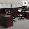 Studio photography of Offices to Go furniture, consisting of two L shape desks against an outside corner wall. There are two chairs and a laptop at each workstation. Overhead is a storage hutch that has silver double doors. This desk uses an American Mahogany (laminate) finish. In the background is another desk for reception, by the window.