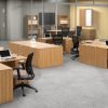 Studio photography of an Offices to Go furniture, set up in an open office near the elevator. Two L shaped desks are pressed against one another, giving a wide surface area for those passing in front. Another set like this is reversed, facing the far back wall. A large credenza exists in the wide area between these work stations. This office's desking uses an Autumn Walnut finish.